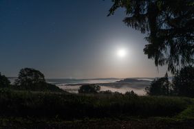 Nebel liegt in einem Tal unter einem sternenklaren Nachthimmel mit hellem Mond, umgeben von Bäumen und Hügeln.