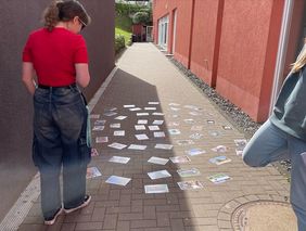 Zwei Personen stehen auf einem gepflasterten Weg zwischen zwei Gebäuden, auf dem zahlreiche Karten oder Bilder verteilt liegen.