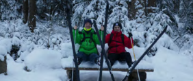 Zwei Personen in Winterkleidung sitzen auf einer Holzbank im Schnee, vor ihnen stehen mehrere Skier im Schnee.